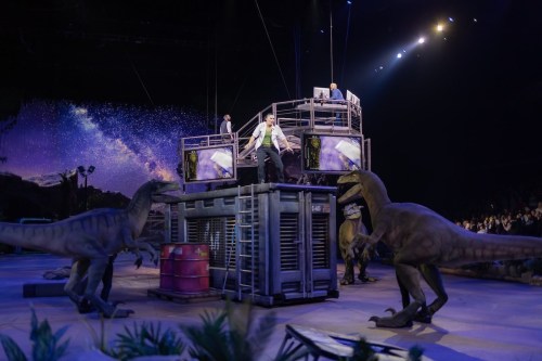 A dramatic scene with actors on a set surrounded by life-sized dinosaur models in a theatrical performance.
