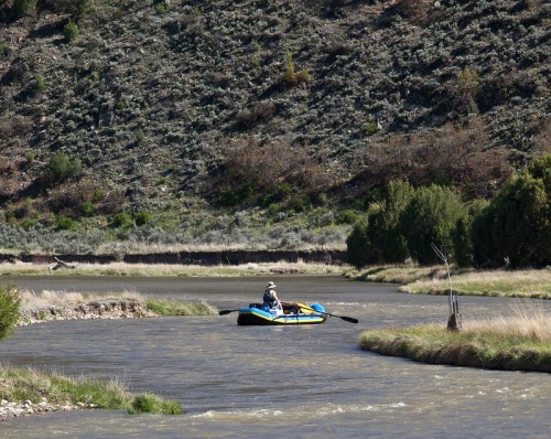 A person paddles a blue raft down a winding river surrounded by green hills and sparse vegetation.