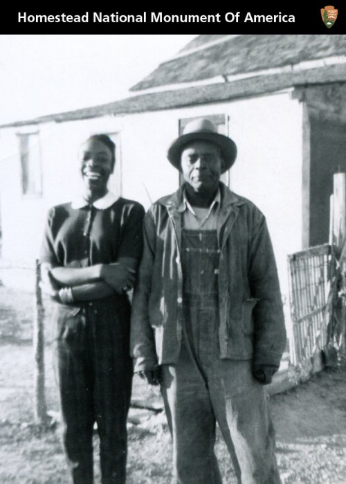 Black and white photo of a man and woman standing together outside a simple house, smiling at the camera.