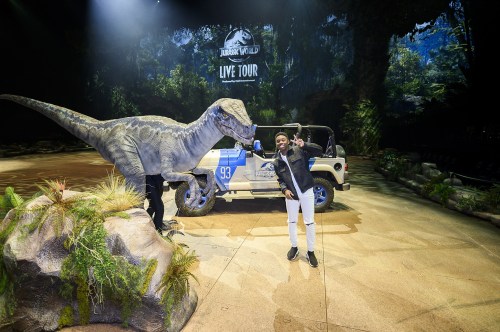 A person poses with a lifelike dinosaur model next to a jeep in a themed Jurassic World live tour setting.