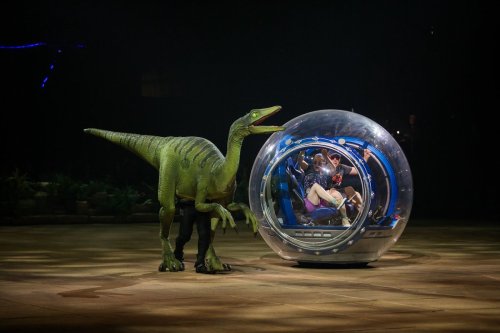 A dinosaur puppet stands next to a futuristic bubble vehicle with two people inside, set in a dark arena.