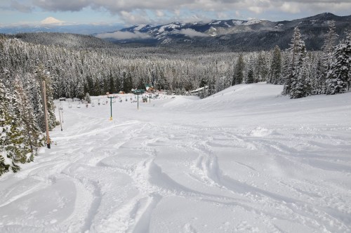 A snowy mountain landscape with ski tracks, trees, and distant mountains under a cloudy sky.