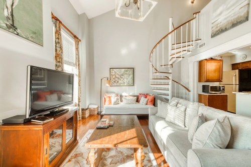 Bright living room with a spiral staircase, cozy seating, a TV, and a kitchen area in the background. Warm, inviting decor.