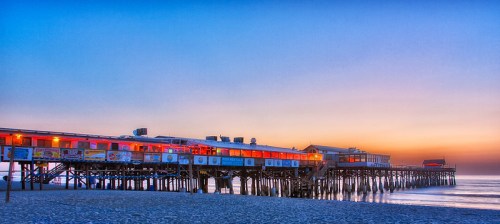 A colorful pier at sunset, with restaurants lining the structure and calm waves lapping at the sandy beach.