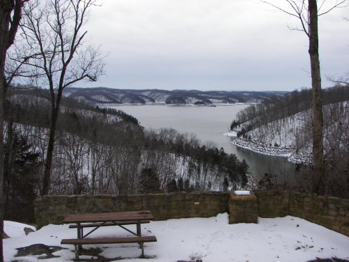 A snowy landscape with a lake surrounded by hills, featuring a stone wall and a picnic table in the foreground.