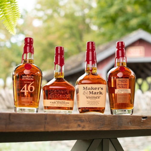Four bottles of Maker's Mark whiskey displayed on a wooden table, with a blurred outdoor background.