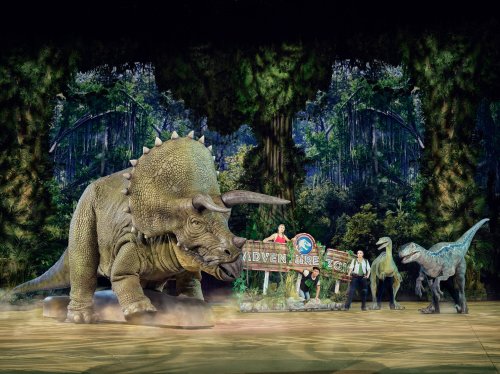 A large triceratops and two smaller dinosaurs on stage with performers in a lush, green forest setting.