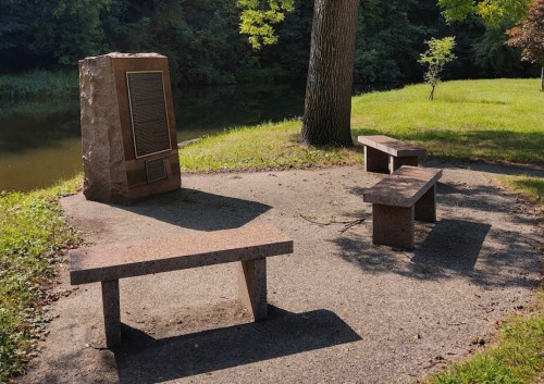 A stone monument with a plaque surrounded by benches near a peaceful pond and trees.