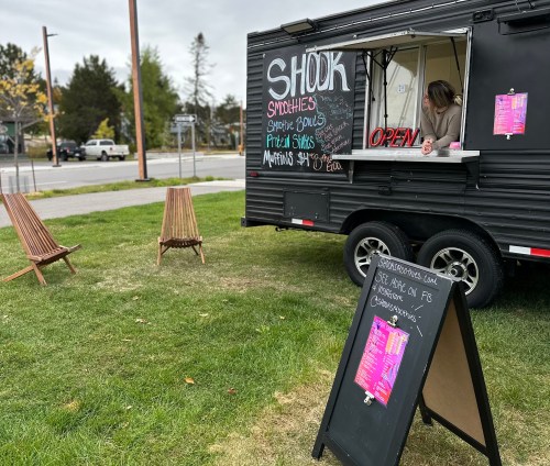 A smoothie food truck with a menu displayed, two wooden chairs nearby, and a sign indicating it's open.