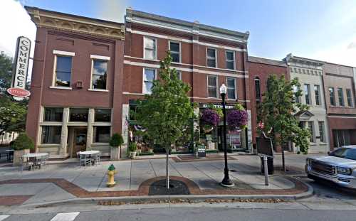 Historic brick buildings line a street, featuring shops, trees, and decorative planters under a clear blue sky.