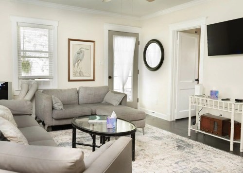 Cozy living room with a gray sectional sofa, round glass table, and light walls, featuring a large mirror and artwork.
