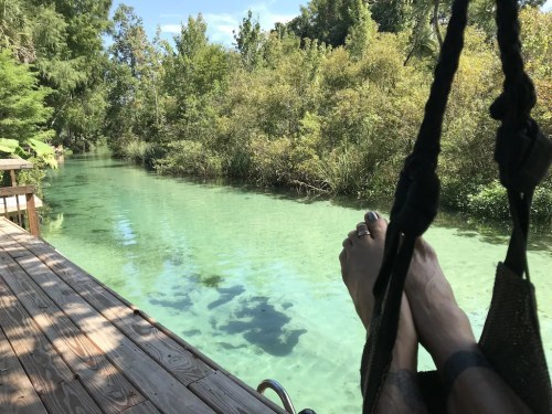 A pair of feet resting on a hammock by a clear, green river surrounded by lush greenery.