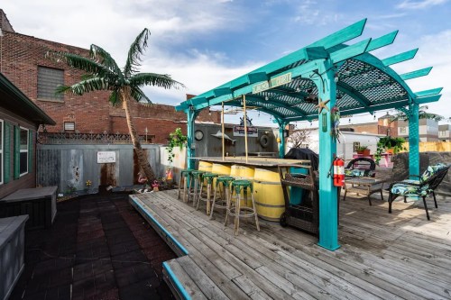 A colorful outdoor bar area with a wooden deck, palm tree, and seating, set against a brick wall backdrop.