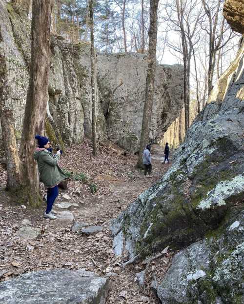 A person takes a photo in a wooded area with large rocks and trees, while two others walk in the background.