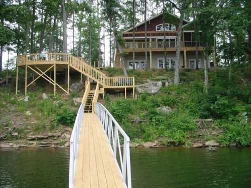 A wooden dock leads to a large house on a hillside, surrounded by trees and overlooking a calm lake.