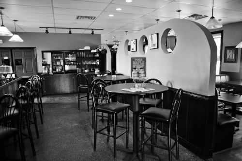 A black and white photo of a cozy café interior with high tables, chairs, and a counter in the background.