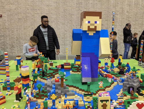 A large LEGO figure stands in a colorful LEGO landscape, with children and adults exploring the creation around it.