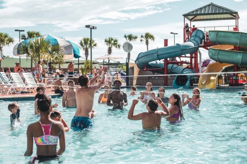 A lively pool scene with children and adults playing and splashing in the water, surrounded by slides and palm trees.