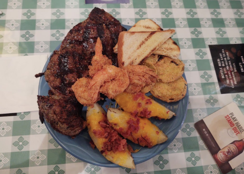 A blue plate with grilled steak, fried shrimp, toasted bread, and stuffed potatoes topped with bacon.