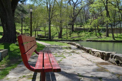 A red bench along a stone path by a calm pond, surrounded by green trees and sunlight.