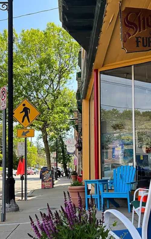 A colorful storefront with blue chairs outside, trees in the background, and a pedestrian crossing sign nearby.