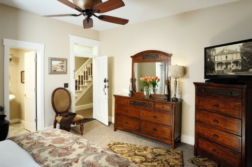 Cozy bedroom featuring a wooden dresser, mirror, TV, and floral decor, with a staircase and bathroom door visible.