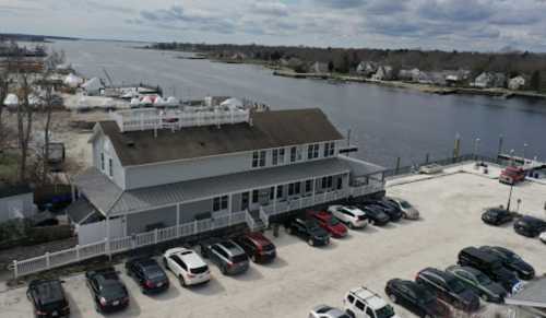 Aerial view of a waterfront building with parked cars, boats in the background, and cloudy skies.