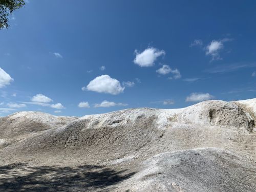 A barren landscape with rolling hills of light-colored soil under a blue sky with fluffy white clouds.