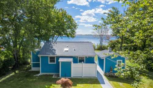 A bright blue house surrounded by trees, with a view of a lake and blue sky in the background.