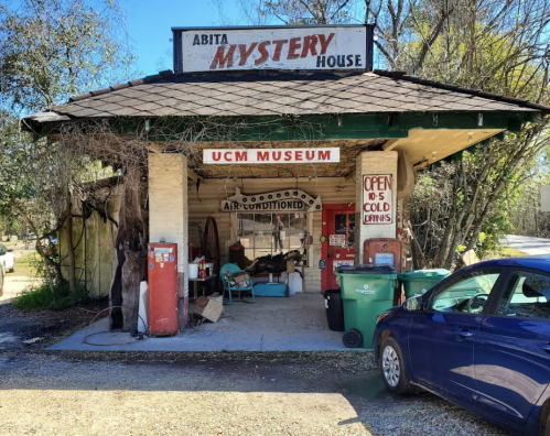 A small, rustic building with a sign reading "Abita Mystery House" and "UCM Museum," featuring vintage gas pumps and a car.