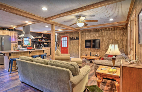 Cozy living room with wooden walls, a ceiling fan, comfortable seating, and a kitchen area in the background.