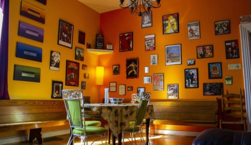 A cozy dining area with an orange wall, adorned with various framed artworks, and a table surrounded by green chairs.