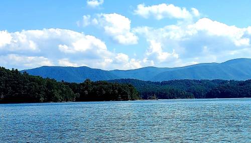 A serene lake surrounded by lush green hills and mountains under a bright blue sky with fluffy white clouds.