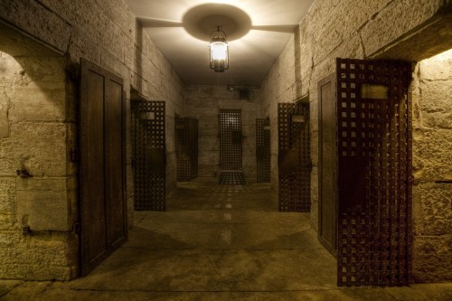 A dimly lit stone corridor with metal doors and a hanging lantern, evoking a historic prison or dungeon atmosphere.