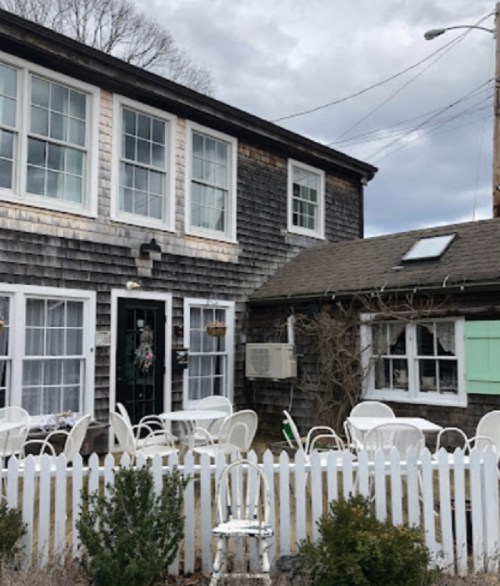 A rustic wooden building with multiple windows, surrounded by a white picket fence and outdoor seating.