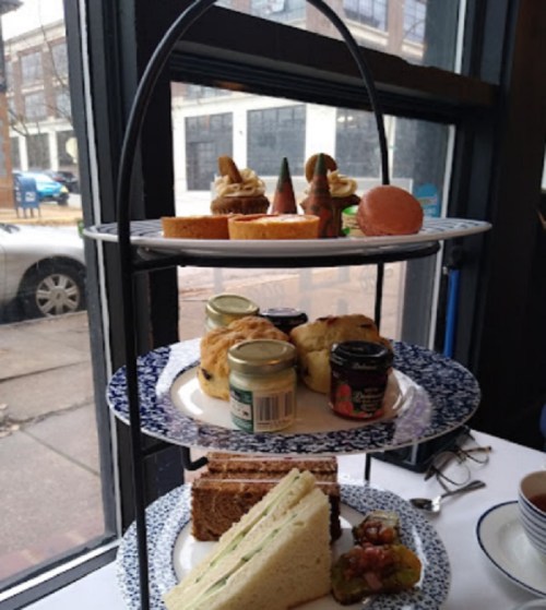 A tiered stand with assorted sandwiches, pastries, and jams, set by a window with a street view outside.