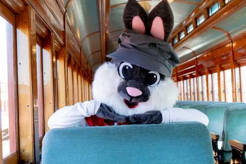 A costumed rabbit character wearing a hat sits on a vintage train, smiling and resting its chin on its hands.