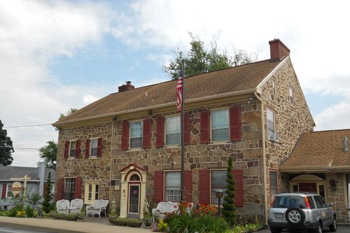 A stone house with red shutters and an American flag, surrounded by greenery and parked cars.