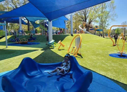 A child sits on a blue play structure in a vibrant playground with slides and climbing equipment under a shaded area.