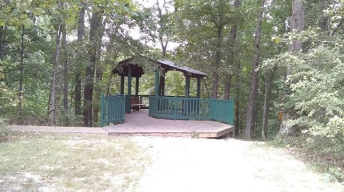 A wooden gazebo surrounded by trees in a forested area, with a deck and seating inside.