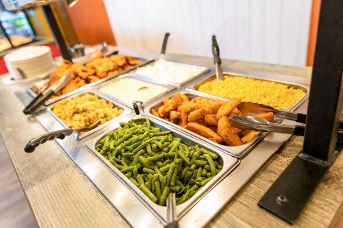 A buffet table with trays of green beans, corn, fried chicken, and sauces, with tongs for serving.