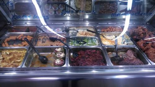 A buffet display featuring various dishes, including meats, sides, and desserts in metal trays with serving utensils.