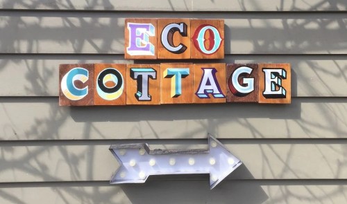 Colorful sign reading "ECO COTTAGE" with an arrow pointing right, mounted on a gray wall.