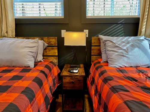 Two twin beds with plaid comforters, a nightstand with a lamp, and sunlight streaming through the windows.