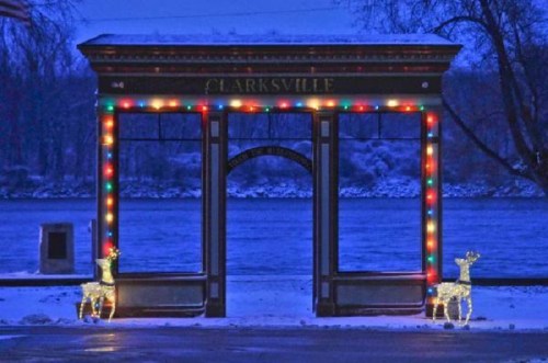 A festive archway decorated with colorful lights and two illuminated reindeer, set against a snowy river backdrop.