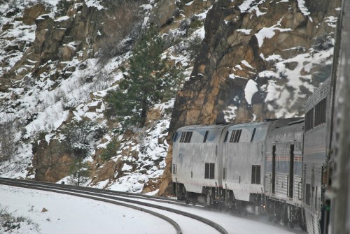 A train curves along snowy tracks beside rocky terrain and pine trees in a winter landscape.