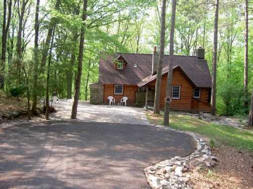 A cozy wooden cabin surrounded by trees, with a gravel driveway and white chairs on the porch.