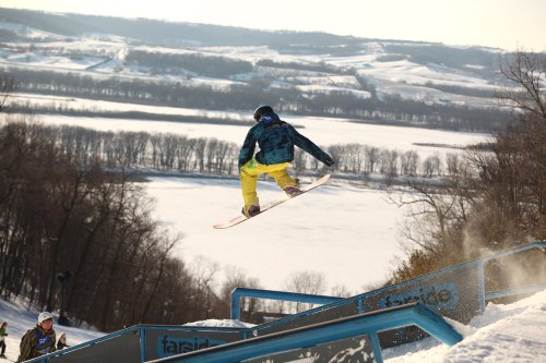 A snowboarder jumps off a rail, soaring over a snowy landscape with trees and a frozen lake in the background.