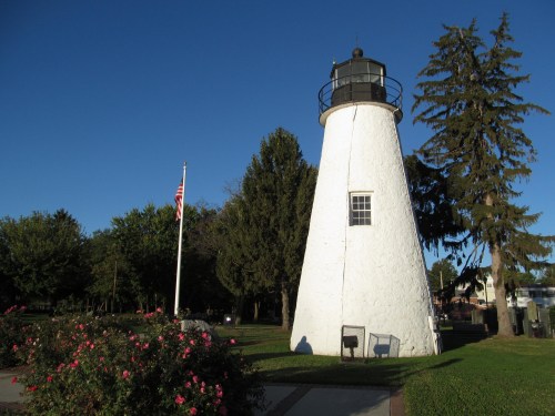 A white lighthouse stands in a park, surrounded by trees and flowers, with a clear blue sky above.