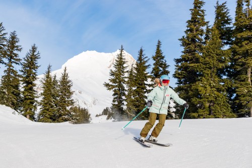 A skier glides down a snowy slope with evergreen trees and a mountain in the background under a clear blue sky.